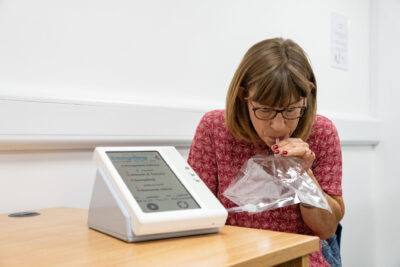 A woman with glasses sits at a table, blowing into a clear plastic bag while a digital device displays sampling instructions nearby.