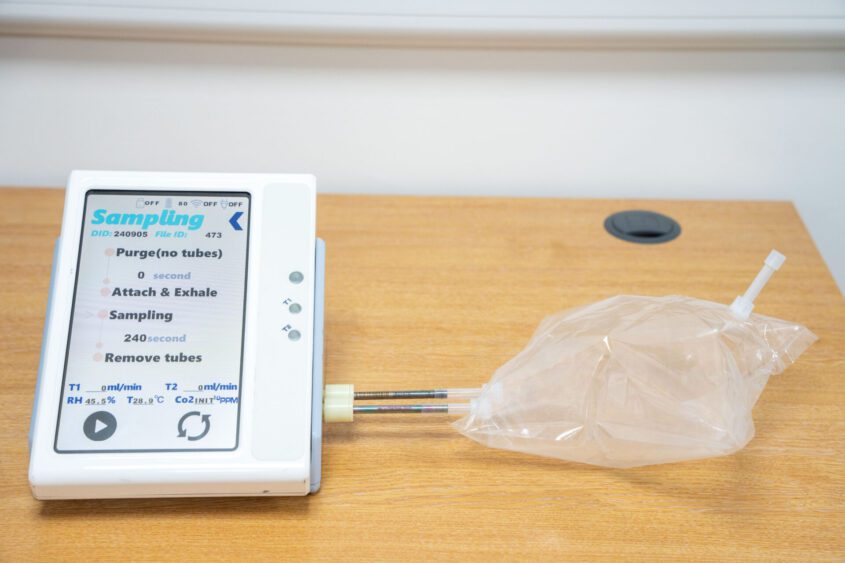 The image shows a medical breathalyser-type device with a touchscreen displaying sampling instructions, connected to a clear plastic bag and a tube, placed on a wooden surface.