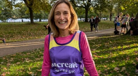 A smiling woman in a purple and yellow running vest stands in a park surrounded by autumn leaves, ready for a race or event.