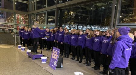 A large group wearing purple sing in front of large glass windows, raising money for Pancreatic Cancer UK