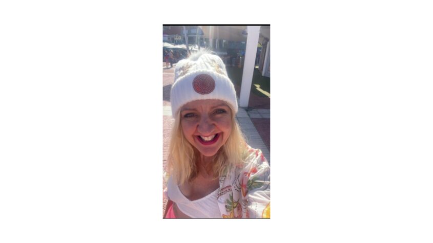 Middle aged woman with blond hair, wearing a white beanie hat and smiling to camera