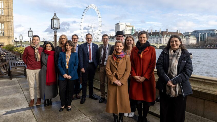 A group of people standing for a picture at the Palace of Westminster (commonly known as the Houses of Parliament), with the London Eye in the background.