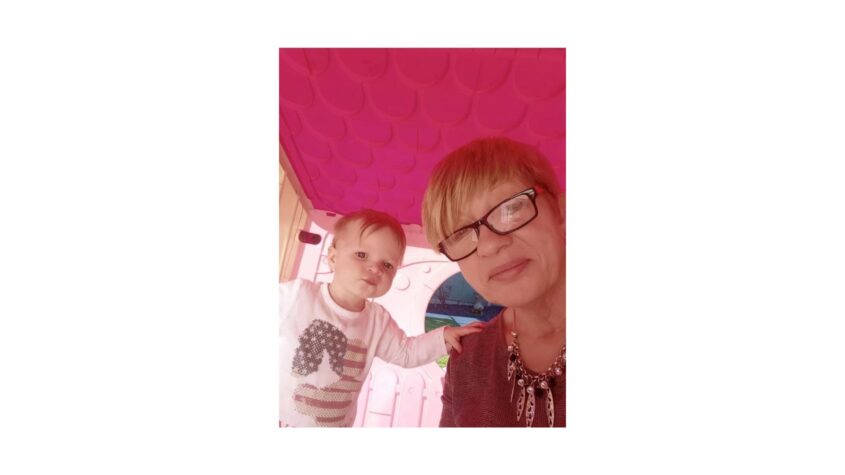 Middle age lady inside a child's playhouse with a bright pink roof, young toddler beside her in the house.