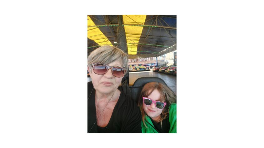 Middle aged lady sitting in a dodgem car at the fairground with a young child beside her, both with sunglasses on.