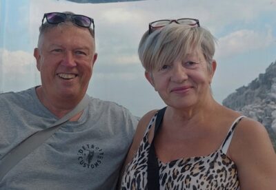 Middle aged man and woman, in a sunny location with blue sky and clouds behind, both smiling to camera