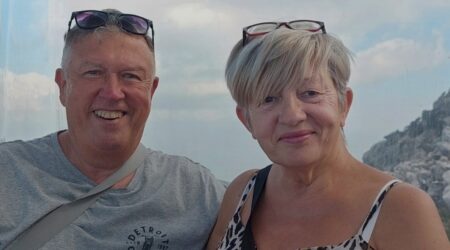 Middle aged man and woman, in a sunny location with blue sky and clouds behind, both smiling to camera
