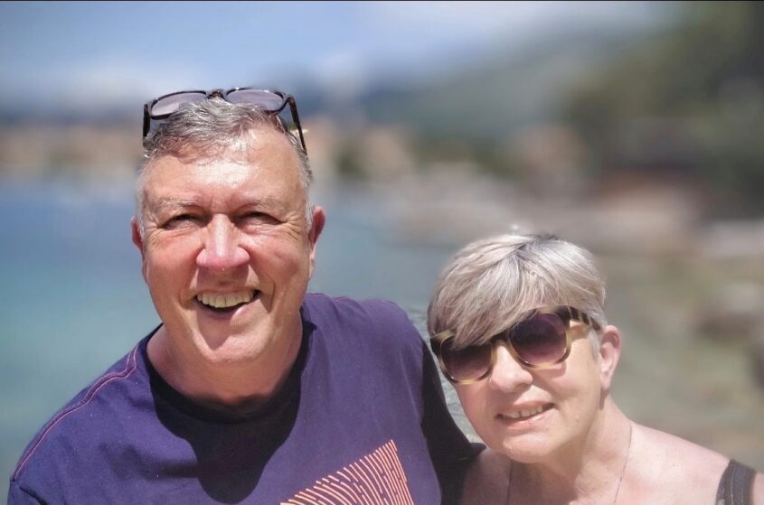 Middle age man and woman in a sunny spot with the sea int he background. both smiling to camera