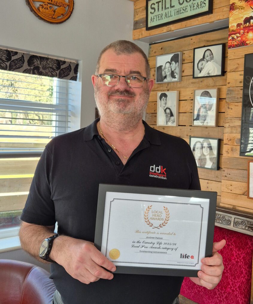 Andrew Palmer smiling, holding his award 'Outstanding Achievement Local Hero' from his local magazine. Behind him hang five photos on a wooden wall of Andrew with Jen
