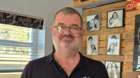 Andrew Palmer smiling, holding his award 'Outstanding Achievement Local Hero' from his local magazine. Behind him hang five photos on a wooden wall of Andrew with Jen