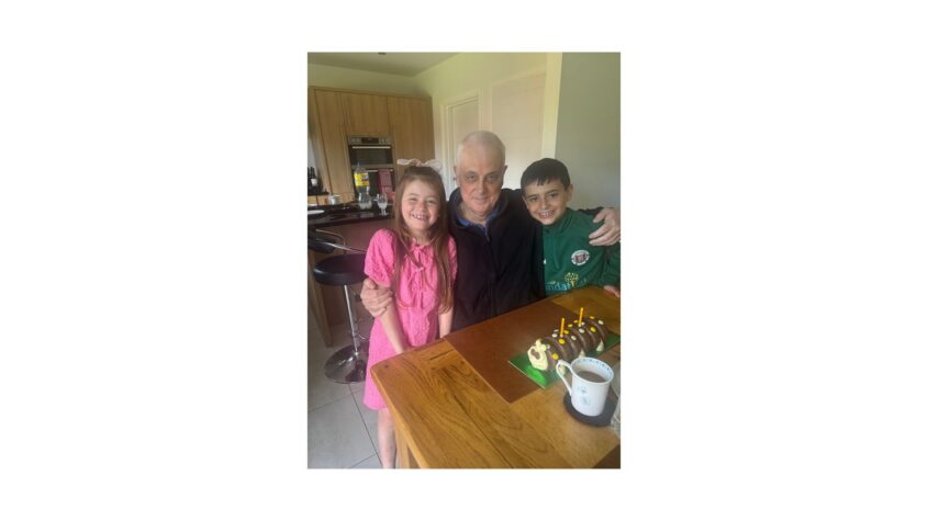 Older man at kitchen table with a chocolate caterpillar cake, two young children standing on either side of him
