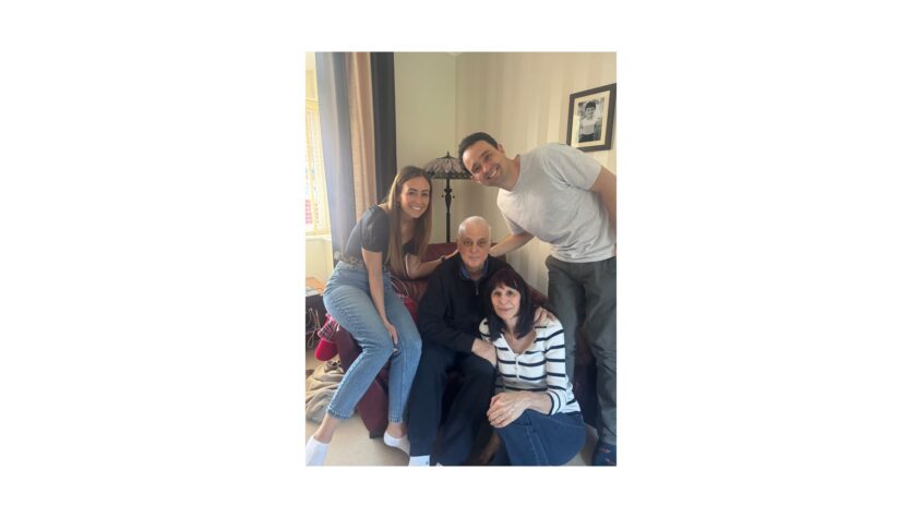 Older man sitting on armchair with younger man and younger woman sitting on either side and older woman sitting on the floor beside him