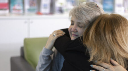 Two women are embracing warmly in a cozy setting, with books and pamphlets visible in the background, suggesting a supportive or comforting environment.