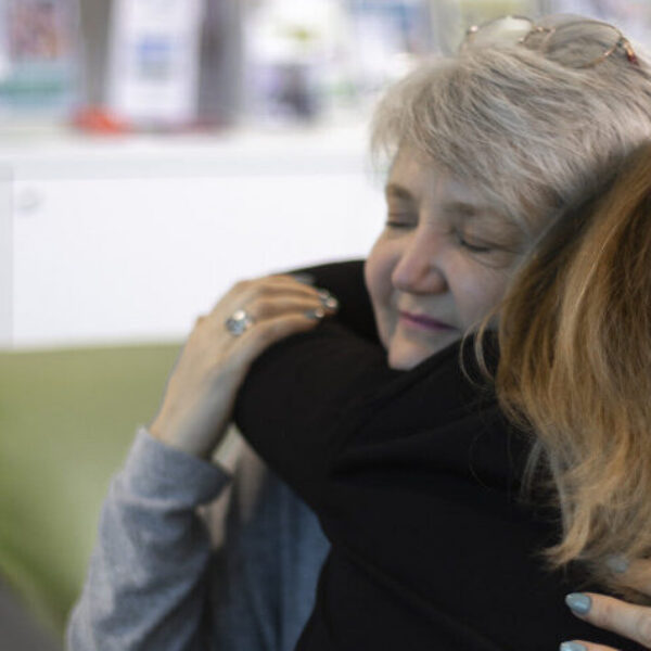 Two women are embracing warmly in a cozy setting, with books and pamphlets visible in the background, suggesting a supportive or comforting environment.