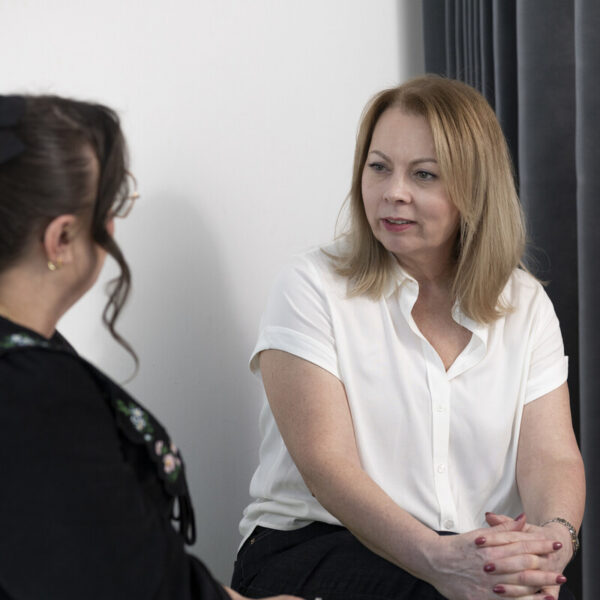 Two women are sitting and talking in a room. One wears a white blouse, the other a black top with a bow in her hair. Gray curtains are in the background.