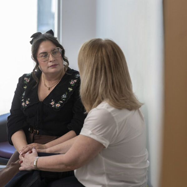 Two women are seated in a room, engaged in conversation. One wears glasses and a black floral top, while the other wears a white shirt. A table is nearby.