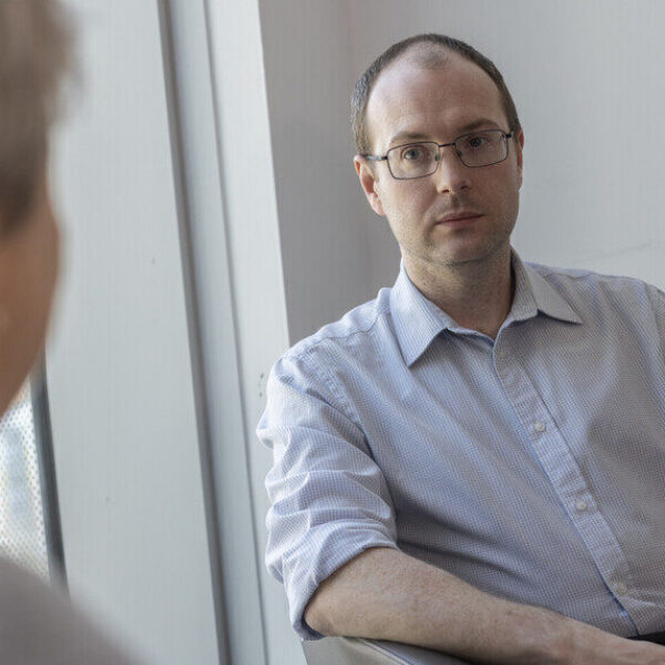 A man in a light shirt sits in a chair, facing a person holding a mug. A book is on the table beside them.
