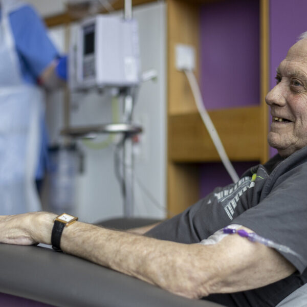 An elderly man smiles while receiving medical treatment in a hospital setting. A healthcare worker in the background wears a blue uniform and apron.