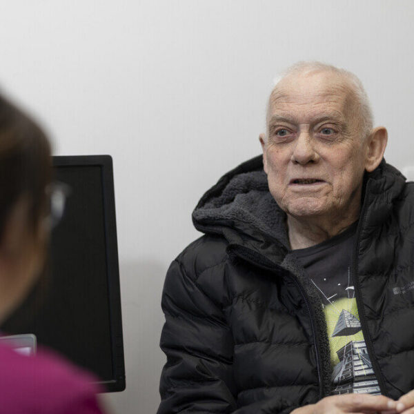 An elderly man in a black jacket sits in a medical office, conversing with a healthcare professional. A computer monitor is visible in the background.