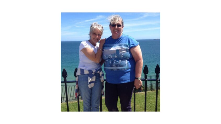 Two woman standing closely together, one with their arm around the other smiling, blue sky and sea in the background