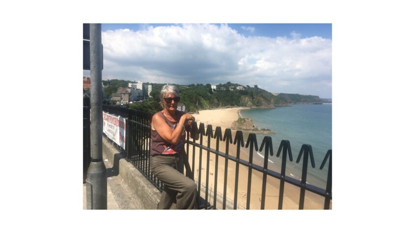 woman wearing sunglasses, sitting beside a low metal fence, overlooking a beach