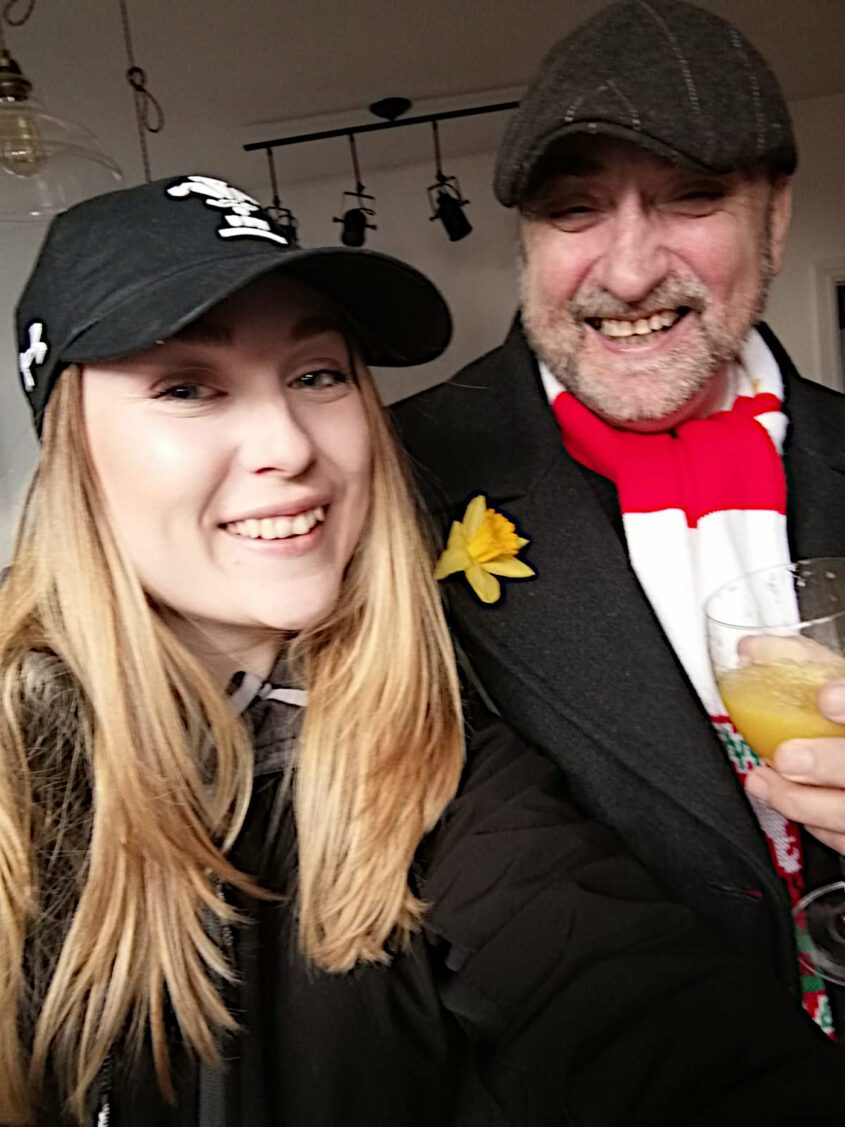 Young woman with long light brown/dark blonde hair, worn down, in black baseball cap. She is smiling and stands next to her dad, who is also smiling and wears a flat cap, daffodil pin and striped red and white scarf.