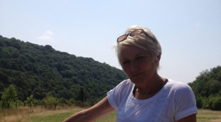 middle aged woman, smiling in the sunshine, sitting on a wooden fence, with fields in the background