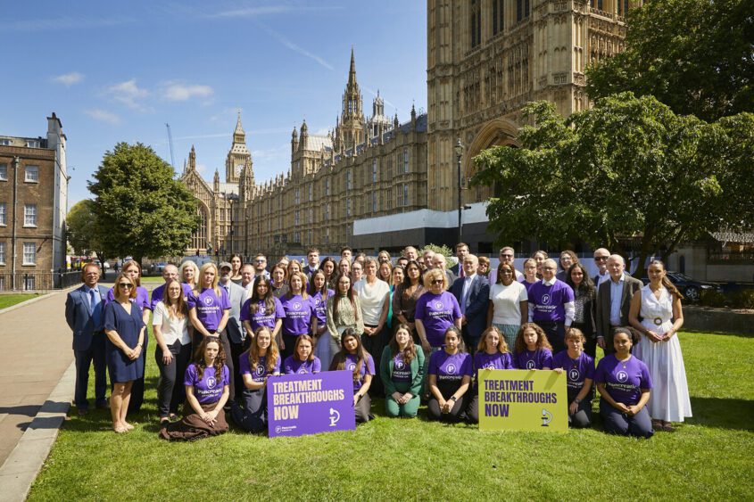 A large group of people posing for a photo outside of the Houses of Parliament. Many are wearing purple Pancreatic Cancer UK t-shirts. Two boards (one purple and one yellow) read 'treatment breakthroughs now'