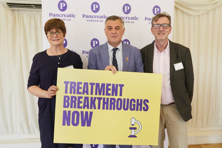 Three people stand for a photo holding a yellow pledge board with 'treatment breakthroughs now' written on it.