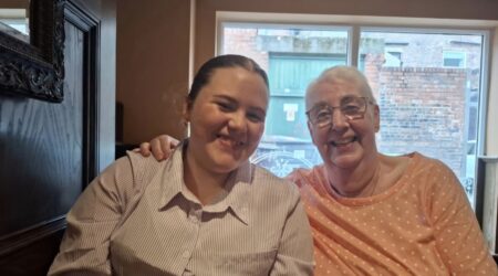 Older woman laughing, with her arm around her granddaughter, both sitting inside a house and looking happy
