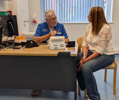 Man in blue hospital scrubs speaks to a woman at a desk in a medical setting. There is a device, the breath test, on the table.
