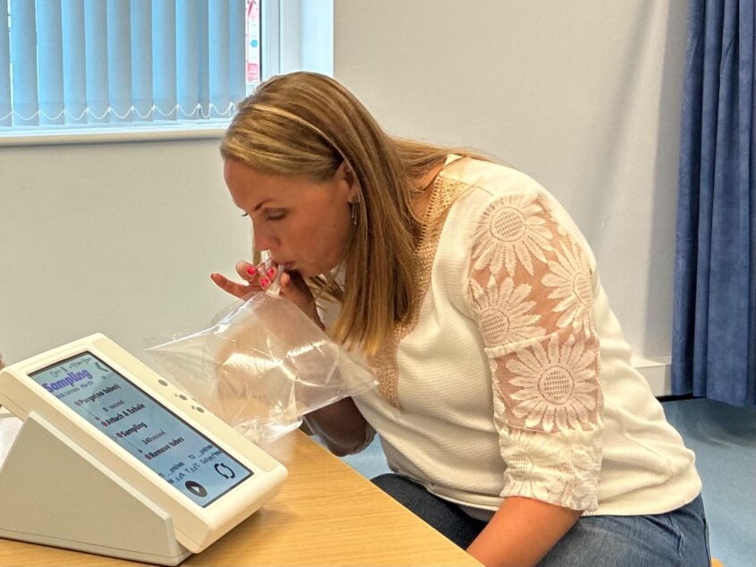 A blonde woman in a GP office, at a desk, breaths into a tube connected to a plastic bag. This is connected to a monitor - the breath test