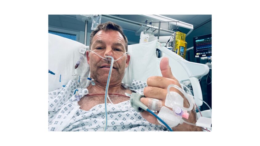 Man in hospital bed with tubes in his nose and hands, giving thumbs up to camera