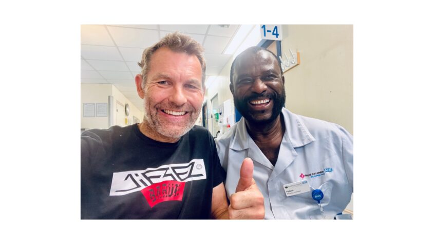 Two men, one in nurse uniform, in hospital corridor. both smiling and the man in black t-shirt giving thumbs up