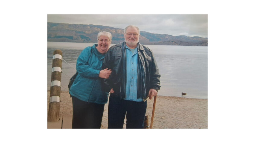 Older man and woman standing on a beach, the sky and water is grey, both are smiling to camera